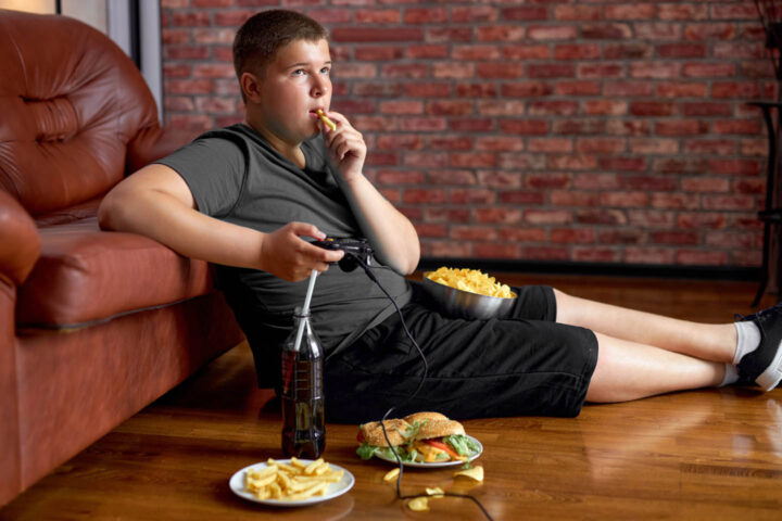 An obese boy eating junk food and playing a videogame in front of the TV